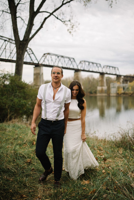 A man in suspenders and a woman in a white dress walk hand in hand near a river, with a railroad bridge and trees in the background on a cloudy day. by Justin Salem Meyer