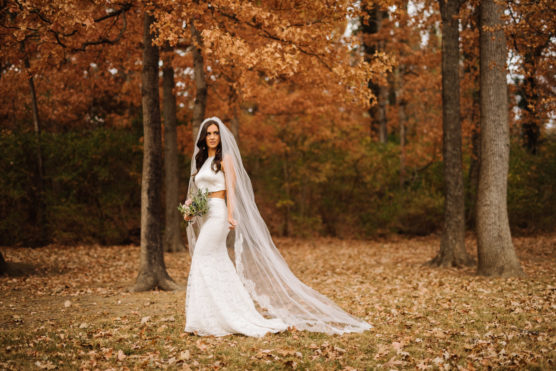 A bride in a white dress and long veil stands outdoors in a forest with autumn leaves, holding a small bouquet and looking at the camera. by Justin Salem Meyer