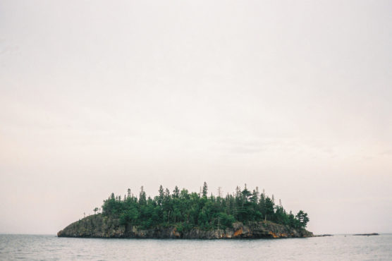 A small, rocky island covered with dense green trees sits in calm water under an overcast sky. The scene appears tranquil and remote, with no visible structures or people. by Justin Salem Meyer
