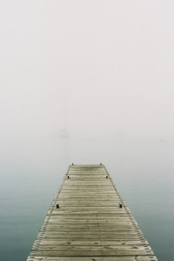 A wooden dock extends over calm, foggy water. Visibility is low due to dense fog, with a faint outline of a sailboat barely visible in the distance. The atmosphere is quiet and serene. by Justin Salem Meyer