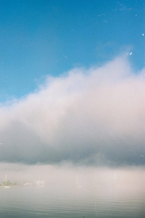 A calm body of water with a thick layer of fog obscuring the horizon; faint outlines of sailboat masts are visible beneath a blue sky with light clouds. by Justin Salem Meyer