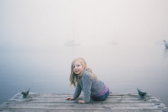 A young girl with long blonde hair crouches on a wooden dock, smiling toward the camera. The background is misty, obscuring the water and boats in the distance, creating a calm and dreamy atmosphere. by Justin Salem Meyer