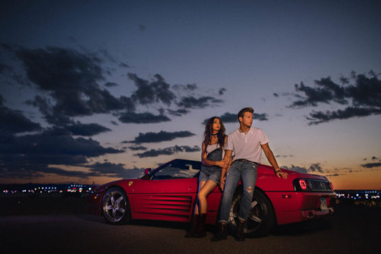 A young man and woman lean against a red convertible sports car at dusk, with a colorful sunset and scattered clouds in the background. The cars headlights are off, and city lights twinkle in the distance. by Justin Salem Meyer