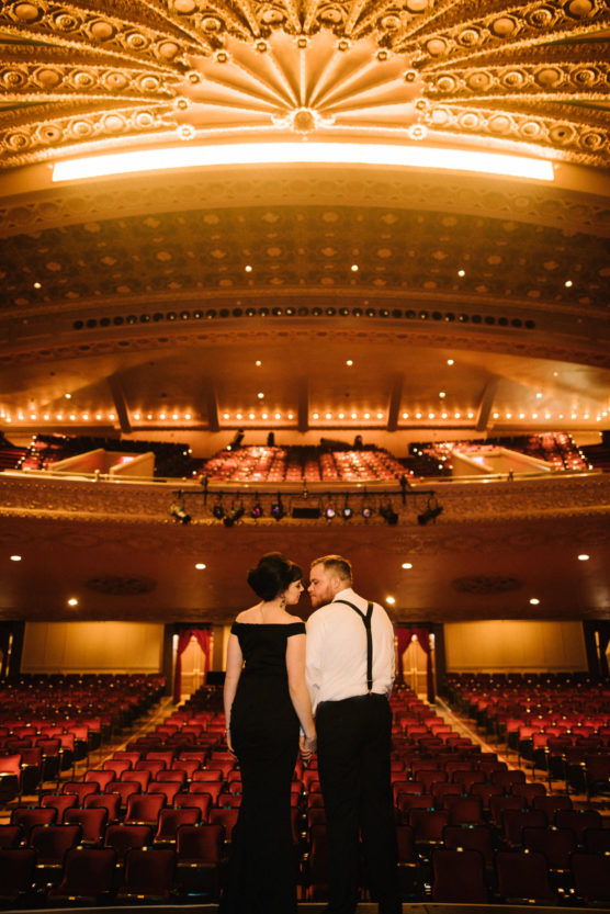 A couple dressed in formal attire stands hand in hand in the center aisle of an ornate, empty theater, gazing at each other beneath an elaborate, illuminated ceiling. by Justin Salem Meyer