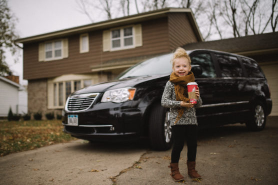 A young child stands on a driveway in front of a black minivan, holding a red cup and smiling. Behind them is a brown two-story house with leafless trees in the background. by Justin Salem Meyer