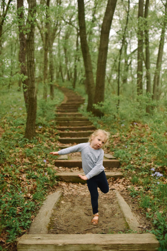 A young child in a gray sweater and navy pants playfully poses on a wooded trail with wooden steps, surrounded by green trees and foliage. by Justin Salem Meyer