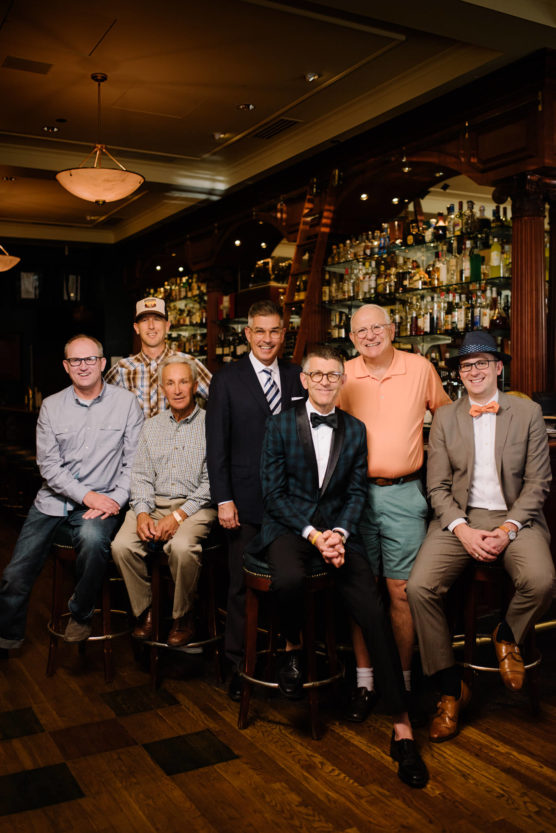 Seven men pose together in a warmly lit bar lined with shelves of bottles. Some are seated on stools while others stand behind, all smiling and dressed in varying styles from casual to formal. by Justin Salem Meyer
