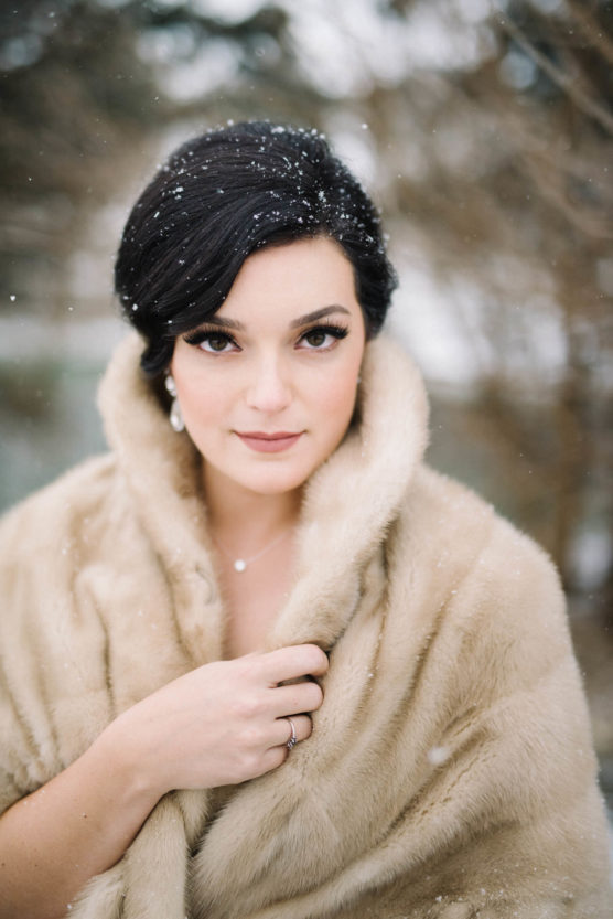 A woman with dark hair styled up stands outside in falling snow, wearing a light brown fur shawl. She gazes at the camera with a soft expression, holding the shawl close around her shoulders. Snowflakes rest on her hair. by Justin Salem Meyer