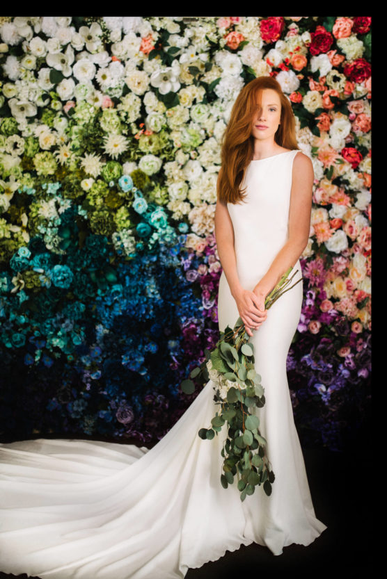 A woman in a white wedding gown holding eucalyptus branches stands in front of a vibrant, multicolored flower wall. Her long red hair is styled to one side, and the dress has a long, flowing train. by Justin Salem Meyer