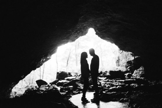 Silhouette of a couple standing face-to-face inside a cave, with bright light streaming in from the cave entrance behind them and rocks scattered on the ground. by Justin Salem Meyer