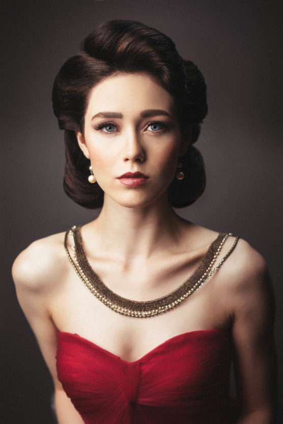 A woman with styled dark brown hair, wearing pearl earrings, a gold necklace, and a strapless red dress, poses against a dark, blurred background, looking directly at the camera with a neutral expression. by Justin Salem Meyer