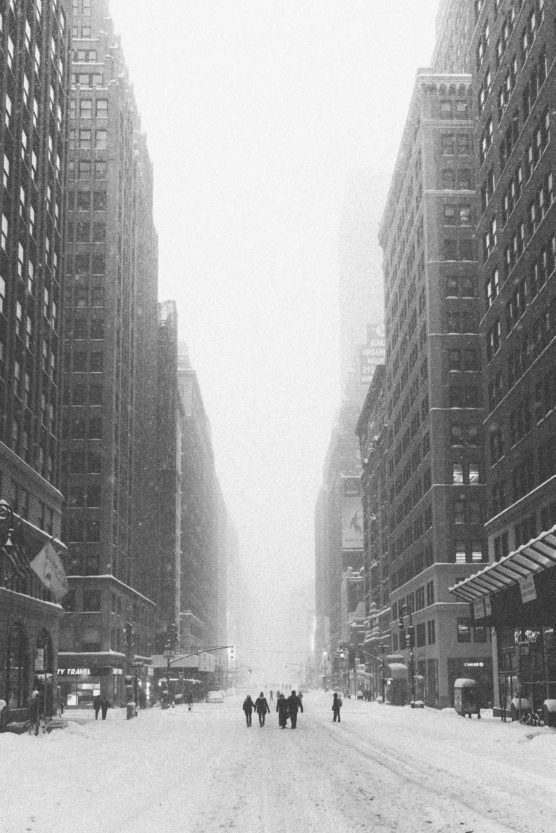 A snowy city street lined with tall buildings, with a few people walking in the middle of the road; the scene is foggy and monochrome, creating a quiet, wintry atmosphere. by Justin Salem Meyer