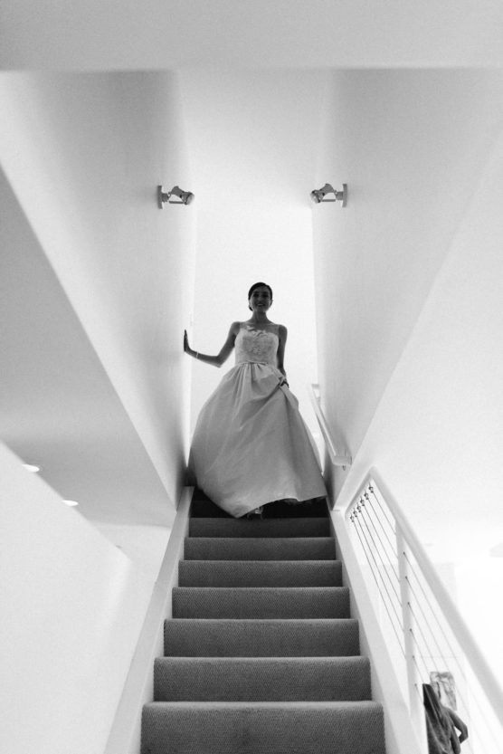 A bride in a wedding gown stands at the top of a carpeted staircase in a modern, bright interior, holding the railing and looking down. The scene is in black and white. by Justin Salem Meyer