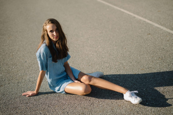 A young woman in a light blue dress and white sneakers sits on an empty asphalt surface, looking toward the camera. Sunlight casts her shadow on the ground. by Justin Salem Meyer