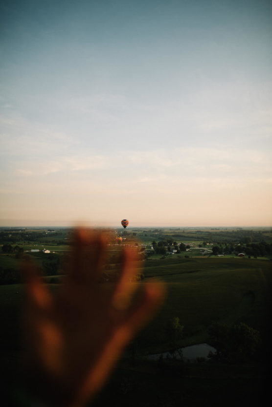 A blurred hand in the foreground reaches toward a distant hot air balloon floating over a scenic countryside landscape at sunset. by Justin Salem Meyer