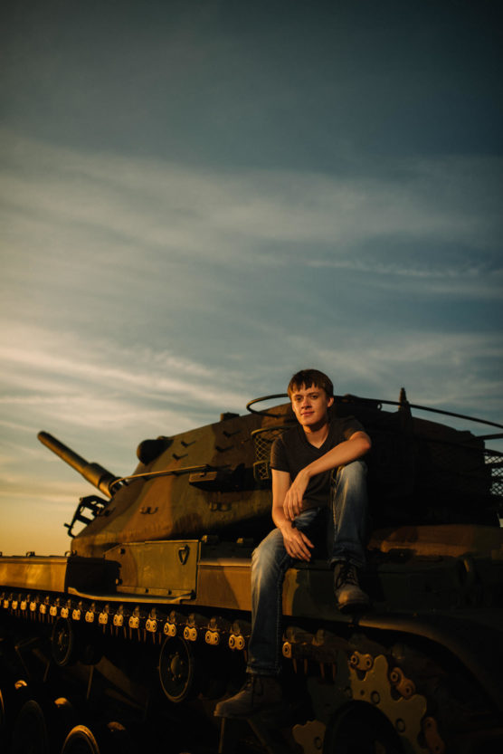 A young man in a dark t-shirt and jeans sits on top of a military tank at sunset, with a calm expression and blue sky in the background. by Justin Salem Meyer