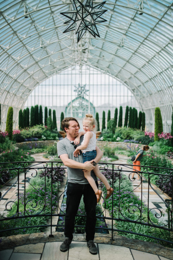 A man holds a young girl in a bright, glass-ceilinged conservatory filled with lush plants and flowers. They stand by a decorative railing, with another person and child visible in the background. by Justin Salem Meyer