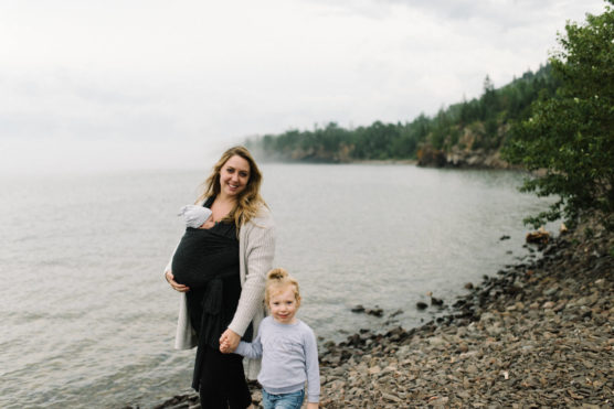 A woman stands on a rocky lakeshore holding a baby in a black carrier and holding hands with a young child, with trees and cloudy sky in the background. by Justin Salem Meyer