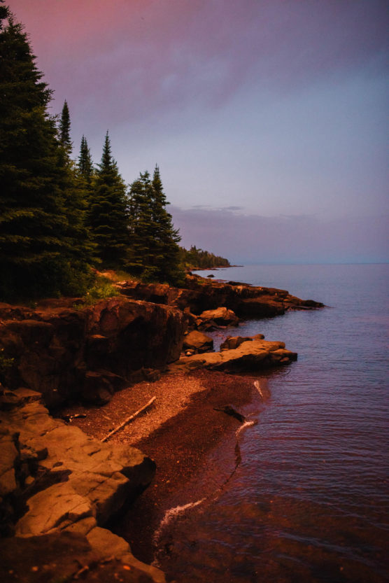 Rocky shoreline with a narrow beach, calm water, and pine trees along the coast at sunset. The sky is tinged with pink and purple hues, creating a peaceful, scenic atmosphere. by Justin Salem Meyer
