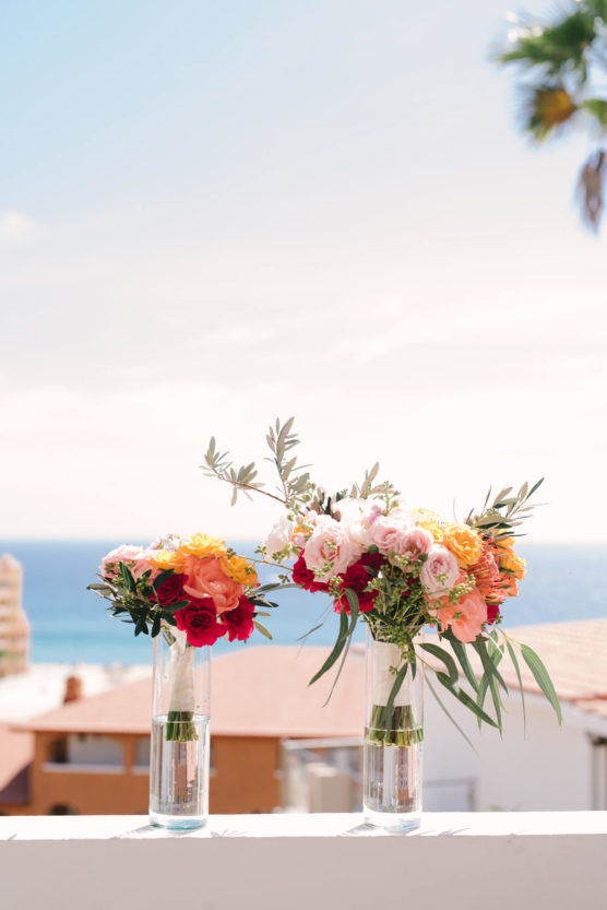 Two glass vases with colorful flower arrangements sit on a white ledge, with a view of rooftops, the ocean, and a partly cloudy sky in the background. by Justin Salem Meyer