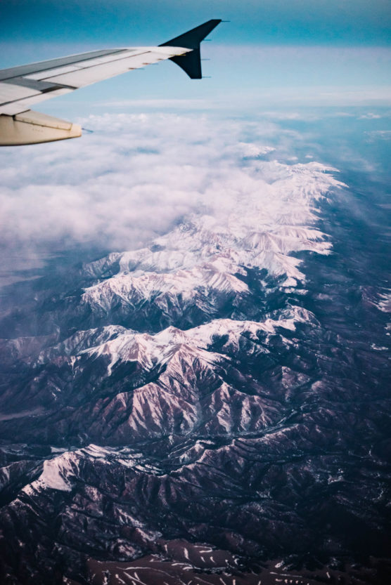 Aerial view of snow-capped mountain range partly covered by clouds, with the wing of an airplane visible in the upper left corner of the image. by Justin Salem Meyer