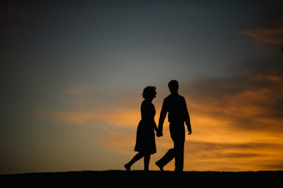 Silhouette of a couple holding hands and walking together against a dramatic sunset sky with orange and blue tones. by Justin Salem Meyer