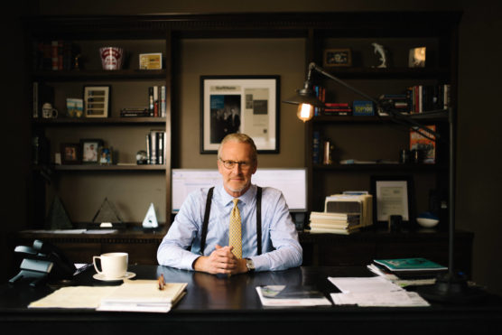 A man in a dress shirt, tie, and suspenders sits at a desk in an office with bookshelves, papers, and a lamp. He faces the camera with his hands folded, and framed items hang on the wall behind him. by Justin Salem Meyer