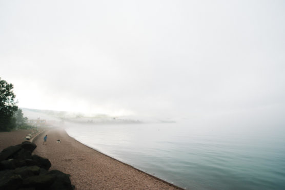 A misty lakeshore with calm water, a person walking a dog along the pebbled beach, and houses partially obscured by fog in the distance. The scene is tranquil and quiet under a gray sky. by Justin Salem Meyer