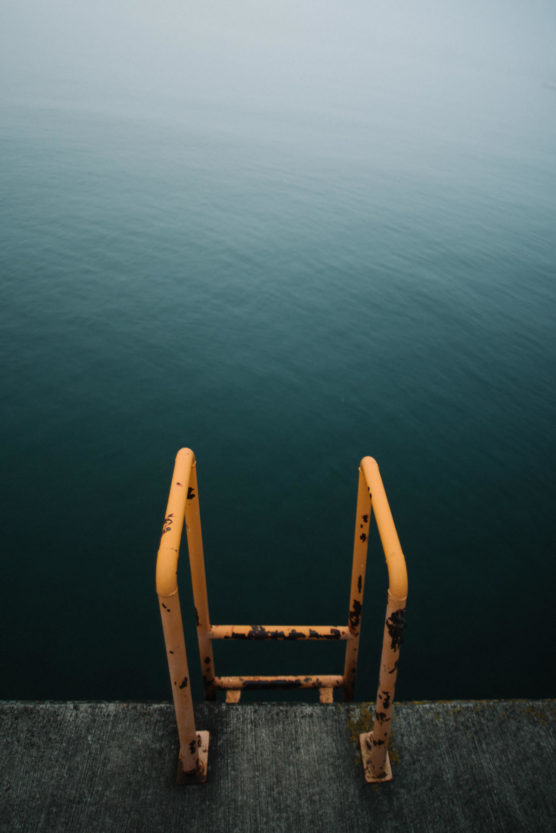 A rusty yellow metal ladder leads down from a concrete platform into calm, blue-green water on a foggy day, with the horizon barely visible in the mist. by Justin Salem Meyer