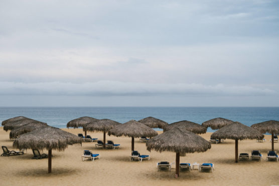 Thatched umbrellas and empty lounge chairs are spread out on a sandy beach, facing a calm blue ocean under a cloudy sky. by Justin Salem Meyer