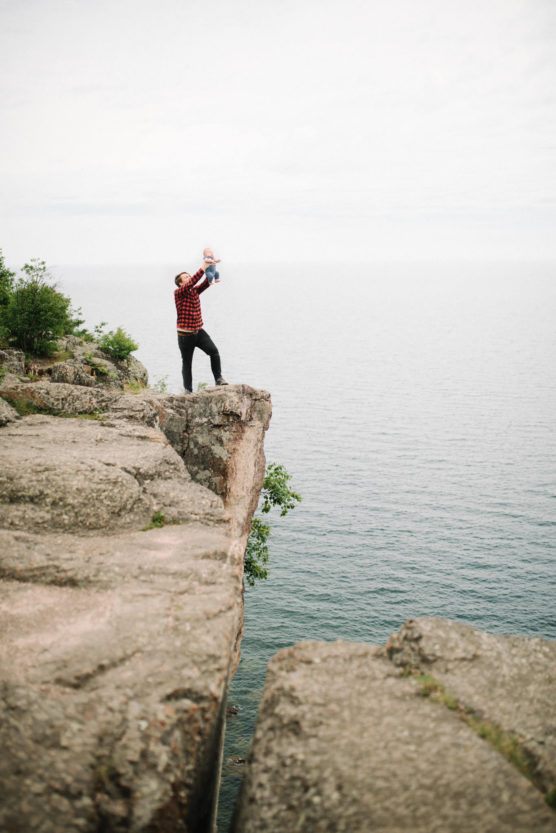 A person in a red plaid shirt stands on the edge of a cliff above a large body of water, lifting a small child joyfully above their head. Trees and rocks surround the cliffside. by Justin Salem Meyer