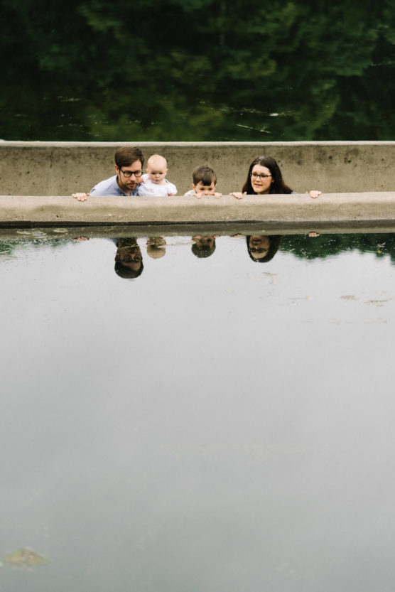 A family of four—two adults and two children—peek over a concrete ledge, with their reflections visible in the still water below. Lush green trees are reflected in the water above them. by Justin Salem Meyer