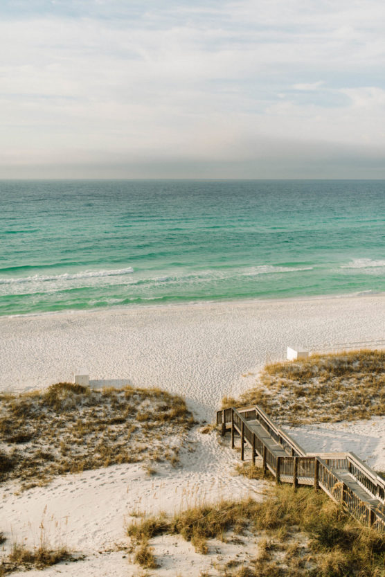 A wooden boardwalk leads over sand dunes to a wide, empty beach with gentle waves and turquoise water under a partly cloudy sky. by Justin Salem Meyer
