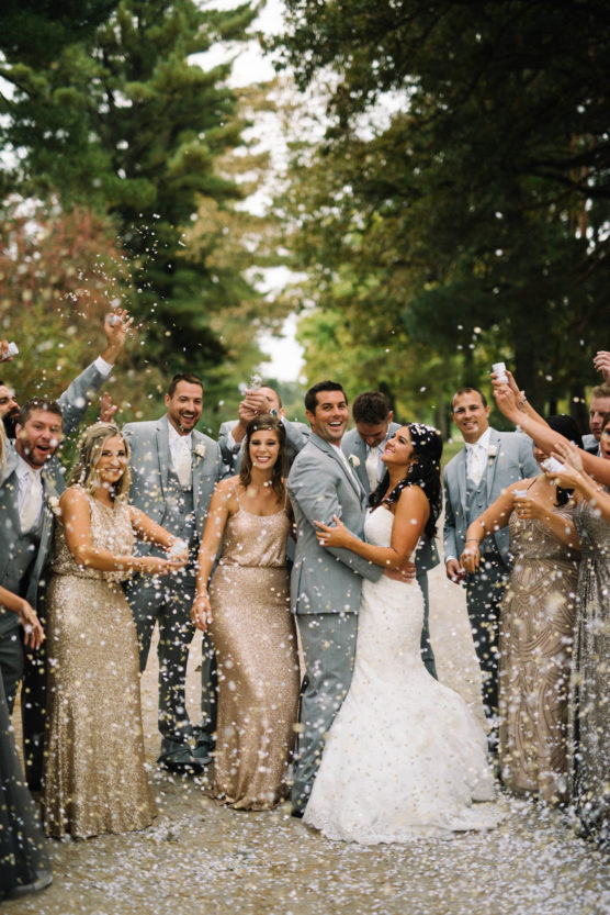 A bride and groom in wedding attire kiss joyfully while surrounded by their cheering wedding party, who are dressed in gold and gray outfits and tossing confetti in the air outdoors. by Justin Salem Meyer