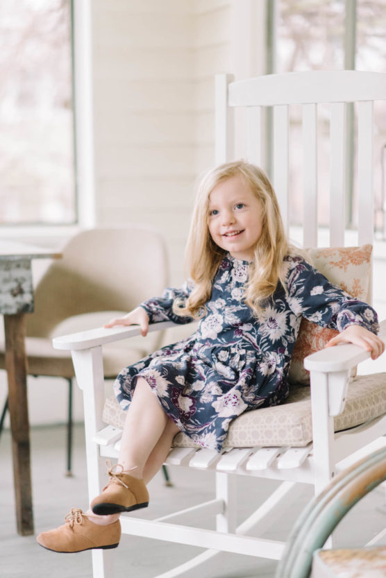 A young girl with long blonde hair, wearing a floral dress and brown shoes, sits smiling on a white rocking chair in a bright room with neutral decor. by Justin Salem Meyer