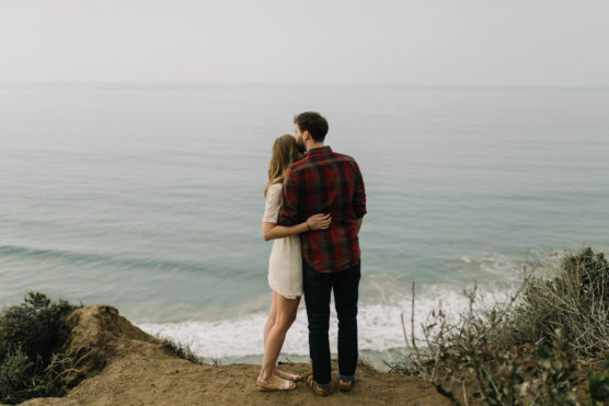 A couple stands on a cliffside overlooking a calm ocean, with their backs to the camera; the woman, wearing a white dress, embraces the man, who is wearing a red plaid shirt and dark jeans. by Justin Salem Meyer