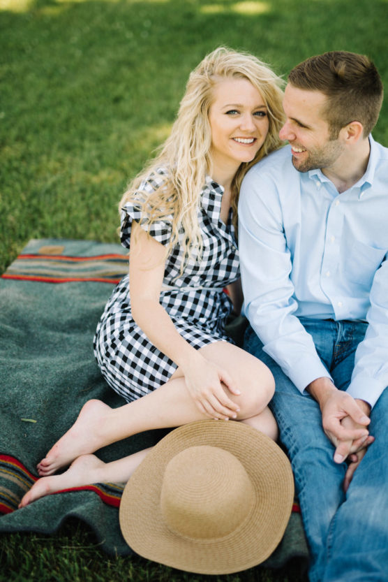 A smiling couple sits closely together on a blanket on the grass. The woman wears a black-and-white checkered dress and holds a sunhat, while the man wears a light blue shirt and jeans. Both look happy and relaxed. by Justin Salem Meyer