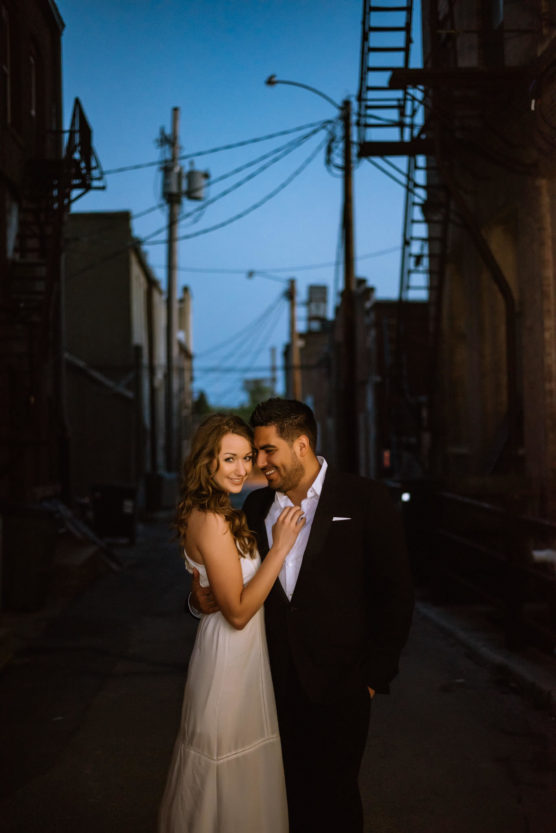 A couple poses closely in a dimly lit urban alley at dusk. The woman in a white dress smiles at the camera, while the man in a black suit looks at her, both surrounded by tall buildings and silhouetted fire escapes. by Justin Salem Meyer