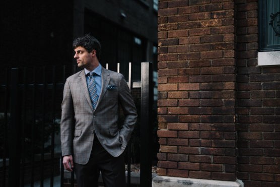 A man in a plaid suit jacket and blue tie stands outdoors by a brick wall and black metal fence, looking to his left with one hand in his pocket. by Justin Salem Meyer