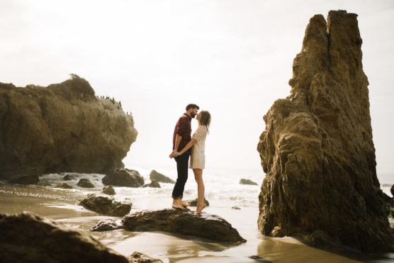 A couple stands barefoot on a rock at the beach, facing each other and holding hands, with large rocky formations and waves in the background under a bright sky. by Justin Salem Meyer