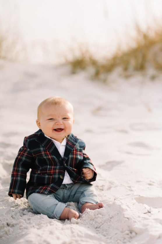 A smiling baby wearing a plaid blazer and jeans sits on white sand with tall grass in the background. by Justin Salem Meyer