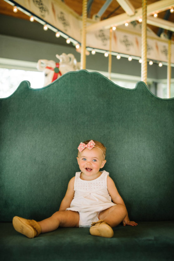 A smiling baby wearing a pink bow headband and light romper sits on a large green bench with a carousel in the background. by Justin Salem Meyer