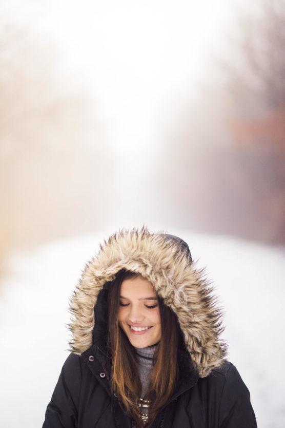 A young woman with long hair smiles while standing outdoors in a snowy landscape, wearing a black coat with a fur-lined hood. The background is softly blurred, creating a bright and serene winter scene. by Justin Salem Meyer