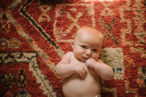 A baby with light skin and reddish-blonde hair lies on a patterned, red and beige rug, looking up with a thoughtful expression and one hand near their mouth. by Justin Salem Meyer