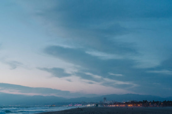 A wide sandy beach at dusk, with gentle waves on the left and a cityscape with glowing lights in the distance under a blue, cloud-filled sky. Mountains are faintly visible on the horizon. by Justin Salem Meyer