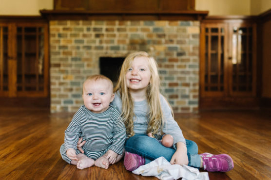 A young girl and a baby sit on a wooden floor in front of a brick fireplace, smiling at the camera. The girl holds a small toy and a blanket; both children appear happy and relaxed. by Justin Salem Meyer