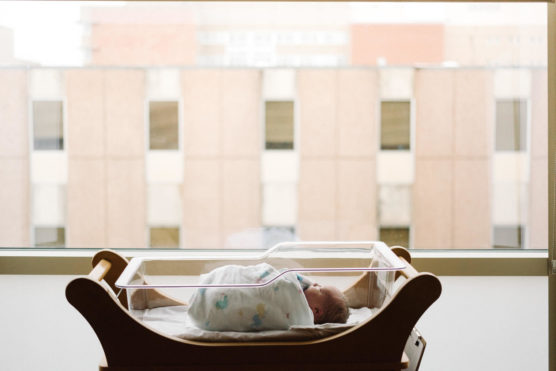A newborn baby wrapped in a blanket lies in a hospital bassinet by a large window, with a blurred view of a multi-story building outside. by Justin Salem Meyer