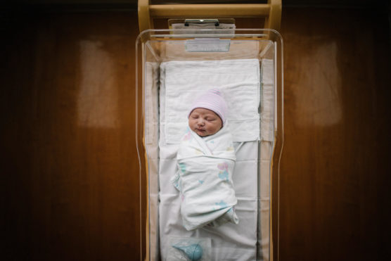 A newborn baby is swaddled in a white blanket and lies in a clear hospital bassinet, wearing a striped hat. The background shows a wooden surface. by Justin Salem Meyer