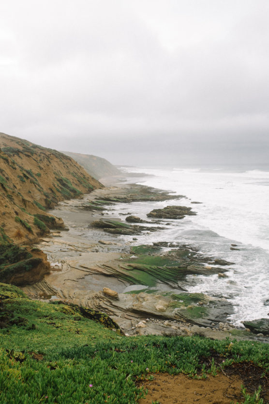 A foggy coastal landscape with rugged cliffs, rocky shoreline, and waves crashing against the shore. Green vegetation grows on the cliff’s edge in the foreground. by Justin Salem Meyer