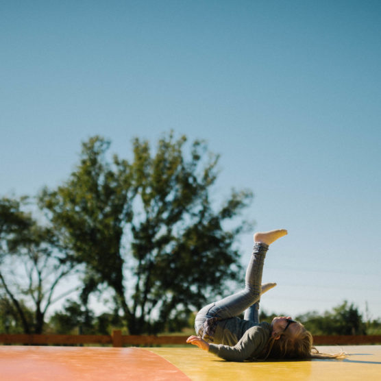 A child lies on their back on a colorful mat outdoors, playfully lifting their legs in the air under a clear blue sky, with trees in the background. by Justin Salem Meyer